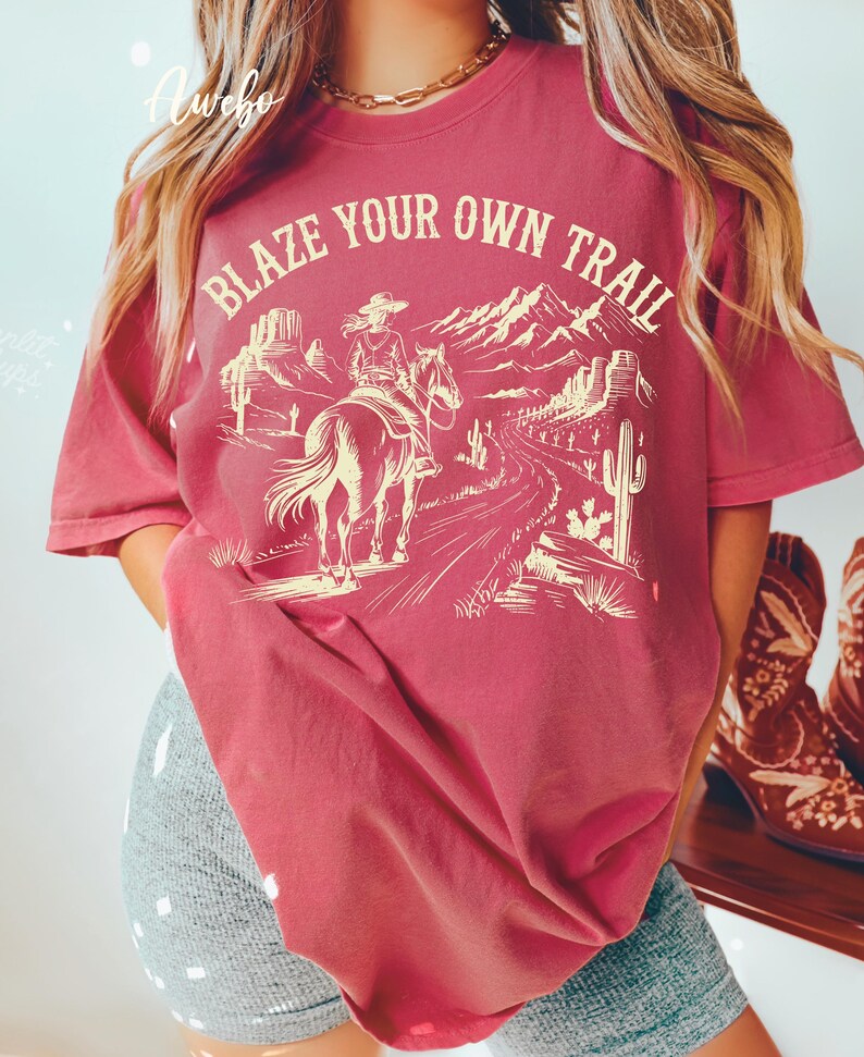 May include: A maroon t-shirt with the text "BLAZE YOUR OWN TRAIL" above a graphic of a person riding a horse through a desert landscape. The design includes mountains, cacti, and a winding road.