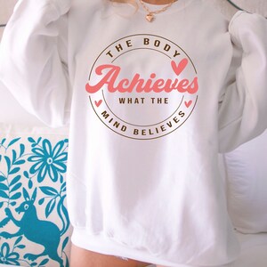 Pode incluir: Camisola branca com um gr&aacute;fico circular castanho que diz "The Body Achieves What The Mind Believes" com cora&ccedil;&otilde;es cor-de-rosa.