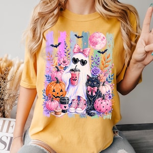 May include: A mustard yellow t-shirt featuring a graphic print of a ghost wearing a pink bow and heart-shaped sunglasses, holding a drink. The design includes a black cat, orange and pink pumpkins, flowers, bats, and a full moon in a colorful setting.