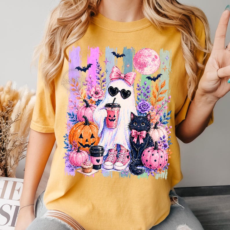 May include: A mustard yellow t-shirt featuring a graphic print of a ghost wearing a pink bow and heart-shaped sunglasses, holding a drink. The design includes a black cat, orange and pink pumpkins, flowers, bats, and a full moon in a colorful setting.