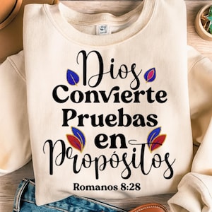 Peut inclure: Sweat-shirt crème avec l'inscription espagnole "Dios Convierte Pruebas en Propósitos" en écriture noire, ornée de motifs de feuilles bleues et rouges. Le texte "Romanos 8:28" est imprimé en dessous. Le sweat-shirt est plié.