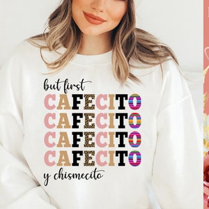 Puede incluir: Sudadera blanca con el texto "but first cafecito y chismecito" en negro y rosa. La palabra "cafecito" se repite cuatro veces con diferentes patrones, incluyendo estampado de leopardo y rayas de colores.
