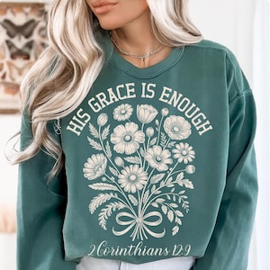 May include: Teal green sweatshirt with the text "HIS GRACE IS ENOUGH" and a floral bouquet design. The text "2 Corinthians 12:9" is printed below the bouquet. The sweatshirt has a crew neck and long sleeves.