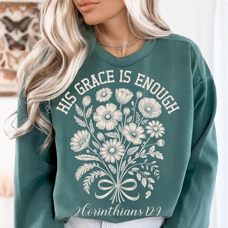May include: Teal green sweatshirt with the text "HIS GRACE IS ENOUGH" and a floral bouquet design. The text "2 Corinthians 12:9" is printed below the bouquet. The sweatshirt has a crew neck and long sleeves.