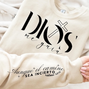 May include: A cream-coloured sweatshirt with the words "DIOS me guia" and a cross graphic in black. The sleeve features the text "Aunque el camino sea incierto Salmo 32:8" in black script. A religious-themed garment.