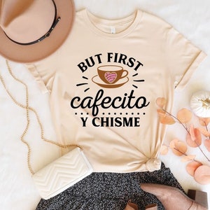 May include: A beige t-shirt with the text "BUT FIRST cafecito Y CHISME" in black. A brown coffee cup with a pink heart is pictured above the text.