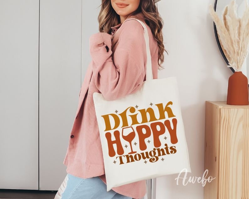Może przedstawiać: Biała torba z pł&oacute;tna z napisem "Drink Happy Thoughts" w pomarańczowych i brązowych literach. Ikona kieliszka do wina jest włączona w tekst. Torbę trzyma osoba w r&oacute;żowym swetrze.