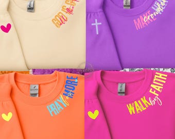May include: Four crewneck sweatshirts in cream, purple, orange, and pink. Each sweatshirt has a different inspirational phrase in colorful text: "God is Good", "Make Heaven Crowded", "Pray More", and "Walk by Faith". Each sleeve has a heart.