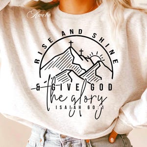 May include: White sweatshirt with a black graphic design featuring a mountain range with crosses on top, a sun, and the text "Rise and Shine & Give God the Glory, Isaiah 60:1".