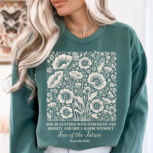 May include: Teal green sweatshirt featuring a floral design with large, stylized flowers and foliage in cream. The text reads "SHE IS CLOTHED WITH STRENGTH AND DIGNITY AND SHE LAUGHS WITHOUT Fear of the Future Proverbs 31:25."