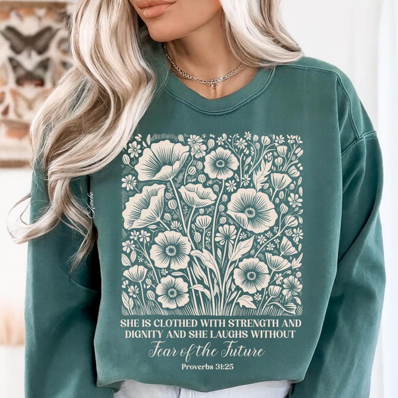 May include: Teal green sweatshirt featuring a floral design with large, stylized flowers and foliage in cream. The text reads "SHE IS CLOTHED WITH STRENGTH AND DIGNITY AND SHE LAUGHS WITHOUT Fear of the Future Proverbs 31:25."