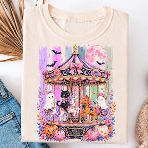 May include: A cream-colored t-shirt featuring a graphic of a carousel with a black cat, a pony, and two ghosts. The design includes a pink moon, bats, and pumpkins at the base. The text "SWEET & SPOOKY" is printed on the carousel base in a dark font.