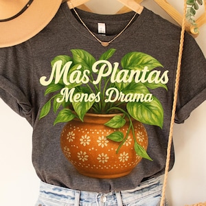 Peut inclure: T-shirt gris foncé avec une illustration colorée d'une plante en pot avec de grandes feuilles vertes. Le pot en terre cuite présente un motif floral. Le texte "Más Plantas Menos Drama" est écrit au-dessus de la plante en vert clair.
