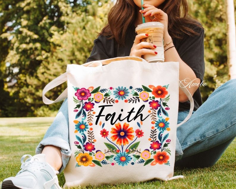 May include: A white canvas tote bag with a floral design and the word "Faith" in black lettering. The bag has a long strap and is being held by a person.