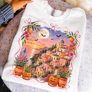 May include: A white crewneck sweatshirt featuring a watercolor-style graphic of a coastal village at sunset. The scene includes a full moon, bats, and a sailboat. The illustration is framed by pink ribbons, orange pumpkins, and colorful flowers.