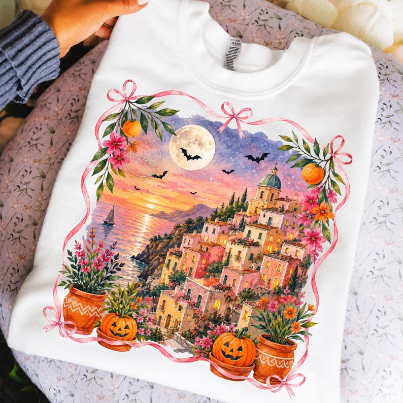 May include: A white crewneck sweatshirt featuring a watercolor-style graphic of a coastal village at sunset. The scene includes a full moon, bats, and a sailboat. The illustration is framed by pink ribbons, orange pumpkins, and colorful flowers.