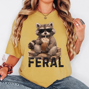 May include: Mustard yellow t-shirt featuring a raccoon wearing sunglasses and drinking a beverage with a straw. The raccoon is sitting on a stack of wooden boxes. The word "FERAL" is printed in black block letters below the raccoon.