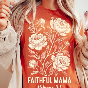 May include: Rust-colored t-shirt featuring a cream-colored floral design with the words "FAITHFUL MAMA" and "Hebrews 11:1" in white script. The shirt has a crew neck and short sleeves.