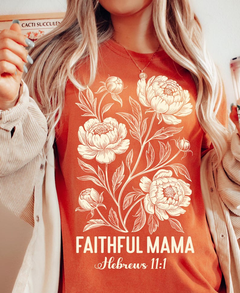 May include: Rust-colored t-shirt featuring a cream-colored floral design with the words "FAITHFUL MAMA" and "Hebrews 11:1" in white script. The shirt has a crew neck and short sleeves.