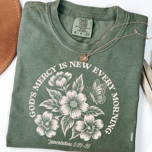 May include: Olive green t-shirt with the text "GOD'S MERCY IS NEW EVERY MORNING" in a circular design with floral and butterfly illustrations. The shirt also features the text "Lamentations 3:22-23". A gold necklace with a pendant rests on the shirt.
