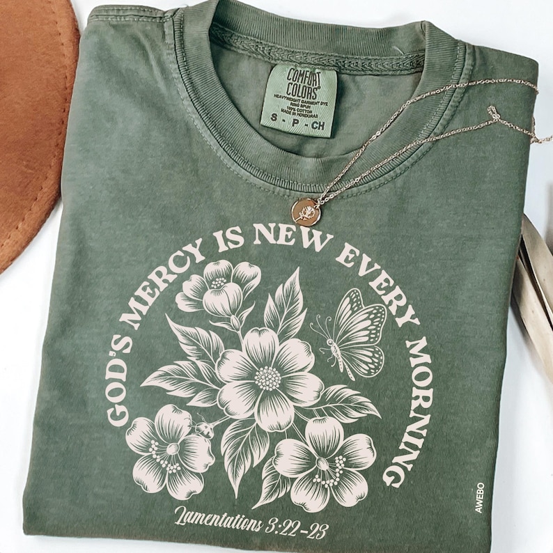 May include: Olive green t-shirt with the text "GOD'S MERCY IS NEW EVERY MORNING" in a circular design with floral and butterfly illustrations. The shirt also features the text "Lamentations 3:22-23". A gold necklace with a pendant rests on the shirt.