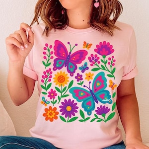 May include: Light pink t-shirt featuring a colorful butterfly and floral design. The graphic includes two large butterflies in pink and teal, surrounded by flowers in various colors. The word "AWEBQ" is printed on the bottom right butterfly.