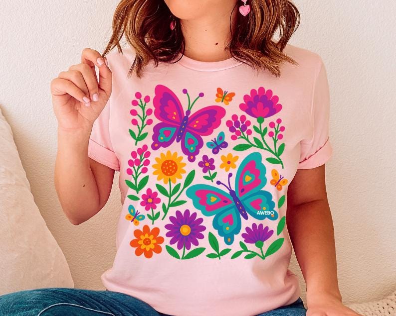 May include: Light pink t-shirt featuring a colorful butterfly and floral design. The graphic includes two large butterflies in pink and teal, surrounded by flowers in various colors. The word "AWEBQ" is printed on the bottom right butterfly.