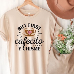 May include: A beige t-shirt with the text "But first cafecito y chisme" in black. The text is surrounded by a decorative design of a coffee cup with a heart-shaped pastry inside.
