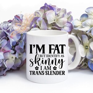 Pu&ograve; includere: Una tazza in ceramica bianca con testo nero che recita "Sono grasso ma mi identifico come magro Sono trans-magro". La tazza &egrave; circondata da fiori viola e bianchi.