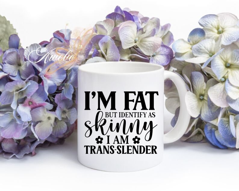 Pu&ograve; includere: Una tazza in ceramica bianca con testo nero che recita "Sono grasso ma mi identifico come magro Sono trans-magro". La tazza &egrave; circondata da fiori viola e bianchi.