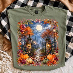 May include: An olive green t-shirt featuring a rectangular graphic of a moonlit cemetery scene at night. The image shows a stone gate, a cobblestone path, autumn trees, and pumpkins surrounded by a floral border with pink ribbons.