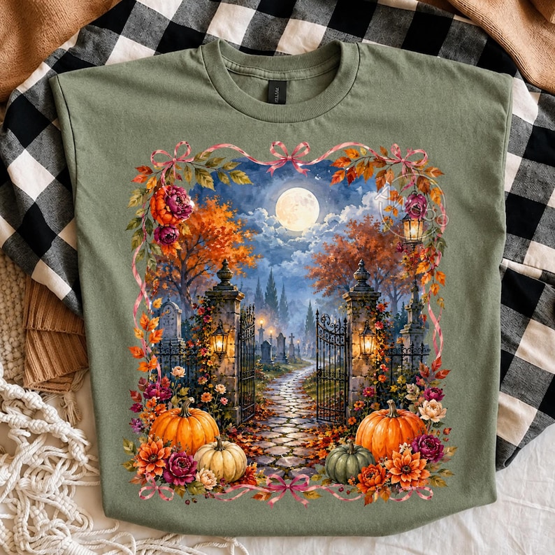 May include: An olive green t-shirt featuring a rectangular graphic of a moonlit cemetery scene at night. The image shows a stone gate, a cobblestone path, autumn trees, and pumpkins surrounded by a floral border with pink ribbons.
