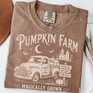 May include: A brown t-shirt featuring a graphic of a vintage truck, a farmhouse, and the text "PUMPKIN FARM" and "MAGICALLY GROWN". The design includes pumpkins, bats, and a crescent moon. The shirt has a Comfort Colors label in the neck area.
