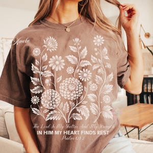 May include: Brown t-shirt featuring a floral design with daisies and large flowers. The text "The Lord Is My Shelter And My Strength, In Him My Heart Finds Rest, Psalm 01-2" is printed below the floral design. The shirt has short sleeves.