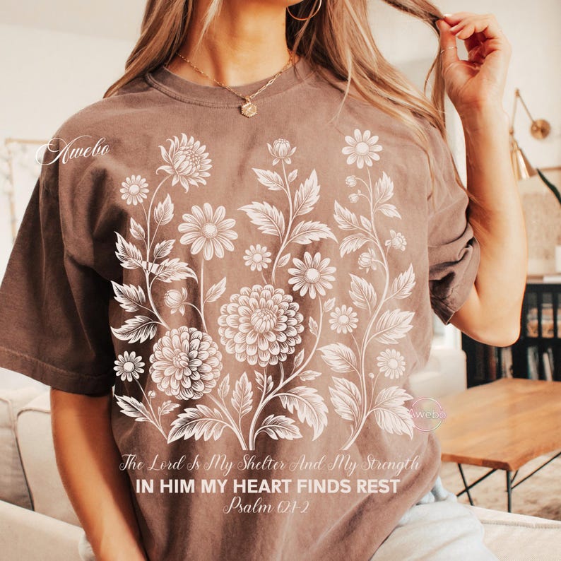 May include: Brown t-shirt featuring a floral design with daisies and large flowers. The text "The Lord Is My Shelter And My Strength, In Him My Heart Finds Rest, Psalm 01-2" is printed below the floral design. The shirt has short sleeves.