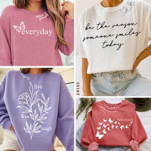 May include: Collection of sweatshirts and t-shirts with inspirational quotes and butterfly graphics. The shirts are in various colors, including pink, white, purple, and red. The text includes "everyday", "be the reason someone smiles today", "enjoy live love laugh", and "create your own sunshine."