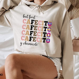 Puede incluir: Una sudadera con capucha beige con el texto "but first cafecito y chismecito" en letras negras y rosas con un estampado de leopardo. Las letras est&aacute;n apiladas una encima de la otra y el texto est&aacute; centrado en la sudadera.