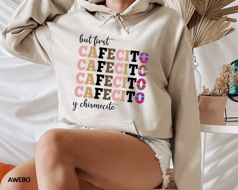 Puede incluir: Una sudadera con capucha beige con el texto "but first cafecito y chismecito" en letras negras y rosas con un estampado de leopardo. Las letras est&aacute;n apiladas una encima de la otra y el texto est&aacute; centrado en la sudadera.