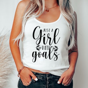 Pode incluir: Camisola branca de al&ccedil;as com texto preto que diz "Just a girl with goals" com um gr&aacute;fico de halteres de cada lado da palavra "with".