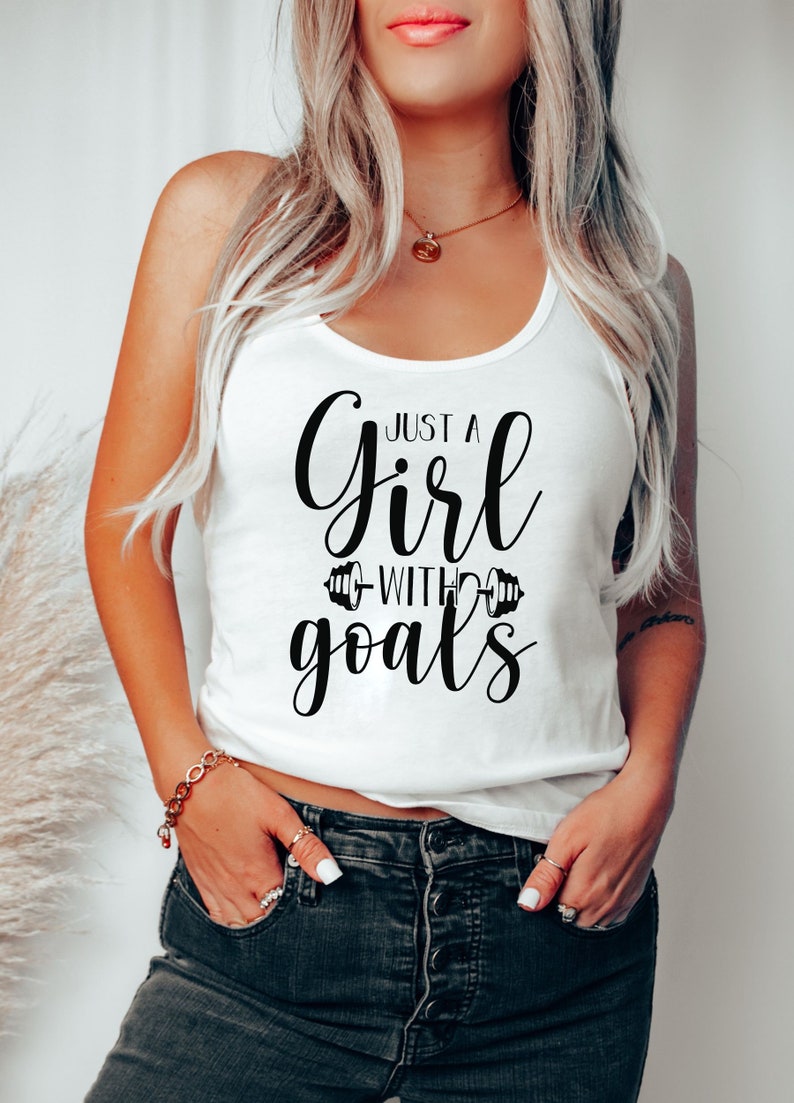 Pode incluir: Camisola branca de al&ccedil;as com texto preto que diz "Just a girl with goals" com um gr&aacute;fico de halteres de cada lado da palavra "with".