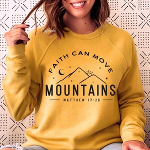 May include: Yellow sweatshirt with a black graphic that says "Faith can move mountains" with a mountain range, moon, and sun graphic. The text "Matthew 17:20" is also included.