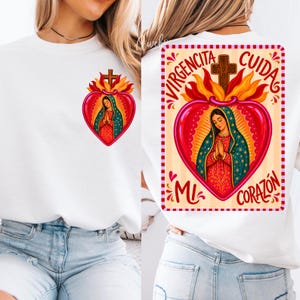May include: A white sweatshirt featuring a graphic of the Virgin Mary inside a red heart with flames and a cross. The back design includes the text VIRGENCITA CUIDA MI CORAZON in red lettering, while the front shows a smaller version of the same heart.