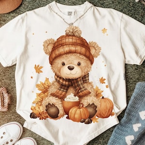 May include: White t-shirt featuring a teddy bear wearing a brown knit hat and plaid scarf. The bear is surrounded by autumn leaves, pumpkins, and a cup of whipped cream. The shirt has a crew neck and short sleeves.