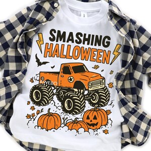 May include: White t-shirt with the text "SMASHING HALLOWEEN" in black and orange. The shirt features an orange monster truck with pumpkins and leaves. A blue and white plaid shirt is draped over the t-shirt.