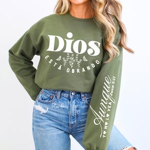 Puede incluir: Sudadera verde oliva con la frase "Dios está Obrando" en blanco, con diseños florales y mariposas. La manga derecha tiene el texto "Aunque Y N LO VEA Juan 5:17" en blanco. La sudadera se combina con vaqueros azules.