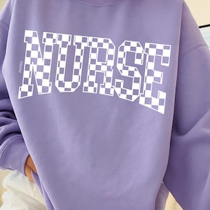 May include: A lilac sweatshirt featuring the word "NURSE" in large, white, chequered letters. The word "AWEBO" is printed vertically on the left side of the sweatshirt.