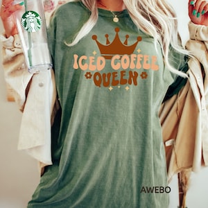 Może przedstawiać: Zielony T-shirt z napisem "Iced Coffee Queen" w brązowych i brzoskwiniowych literach z brązową koroną nad tekstem. Koszulka ma postarzony wygląd, a tekst ma styl retro.