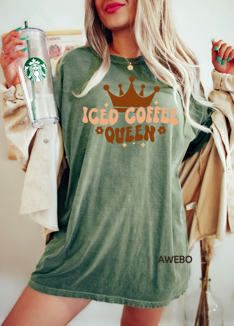 Może przedstawiać: Zielony T-shirt z napisem "Iced Coffee Queen" w brązowych i brzoskwiniowych literach z brązową koroną nad tekstem. Koszulka ma postarzony wygląd, a tekst ma styl retro.