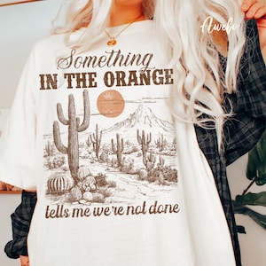 May include: Cream-colored t-shirt with a desert landscape graphic. The graphic features cacti, a sun, and mountains in brown tones. Text on the shirt reads "Something in the Orange tells me we're not done."