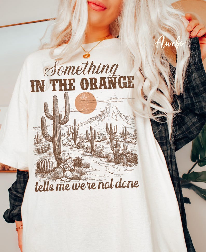 May include: Cream-colored t-shirt with a desert landscape graphic. The graphic features cacti, a sun, and mountains in brown tones. Text on the shirt reads "Something in the Orange tells me we're not done."